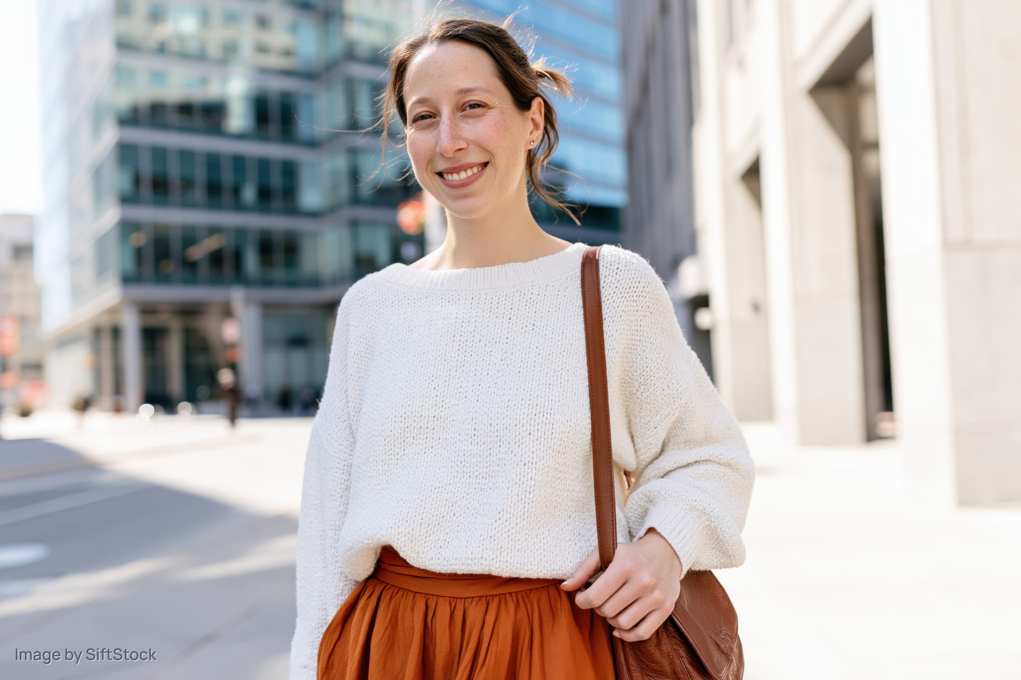 Woman smiling on city sidewalk wearing white sweater and brown skirt with leather handbag