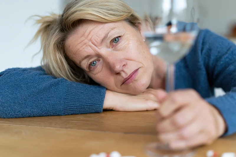 Middleaged woman resting her head on a wooden table while holding a wine glass, looking sad and tired
