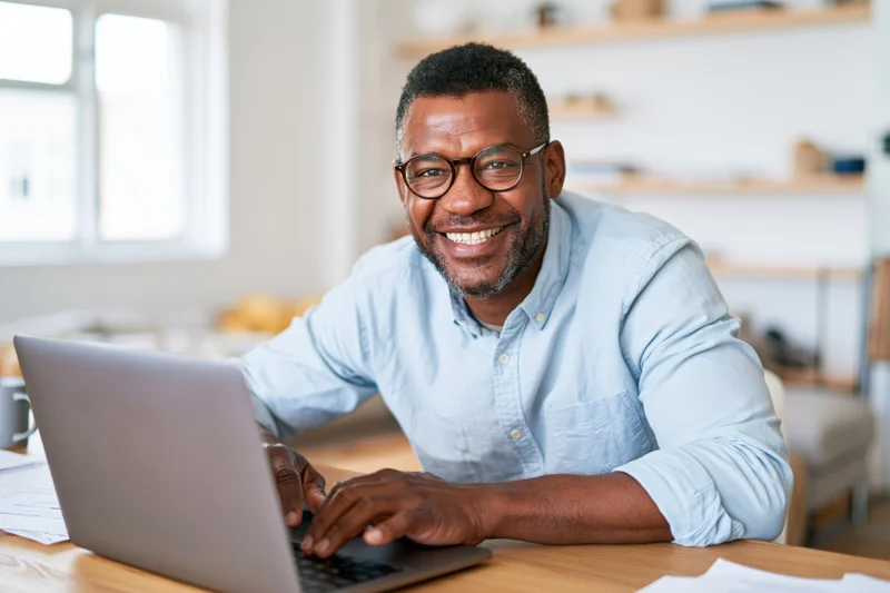 Man smiling while typing on laptop at wooden desk in home office
