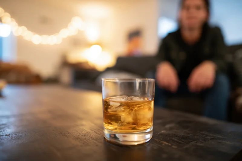 Whiskey with ice in a short glass on a dark wooden table with a woman sitting blurred in the background
