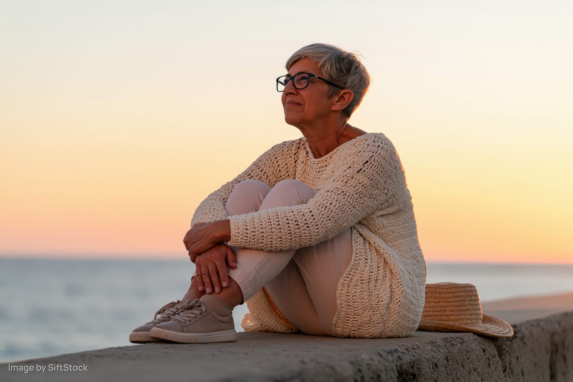 Senior woman sitting on concrete ledge by ocean at sunset, wearing knit sweater and glasses, hat beside her