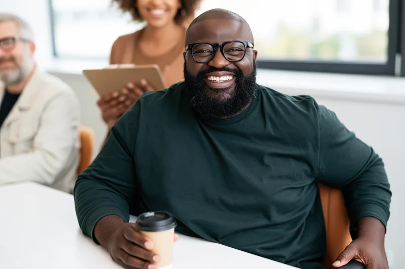 Bearded man with glasses smiling while holding coffee cup at white desk in office meeting with colleagues in background