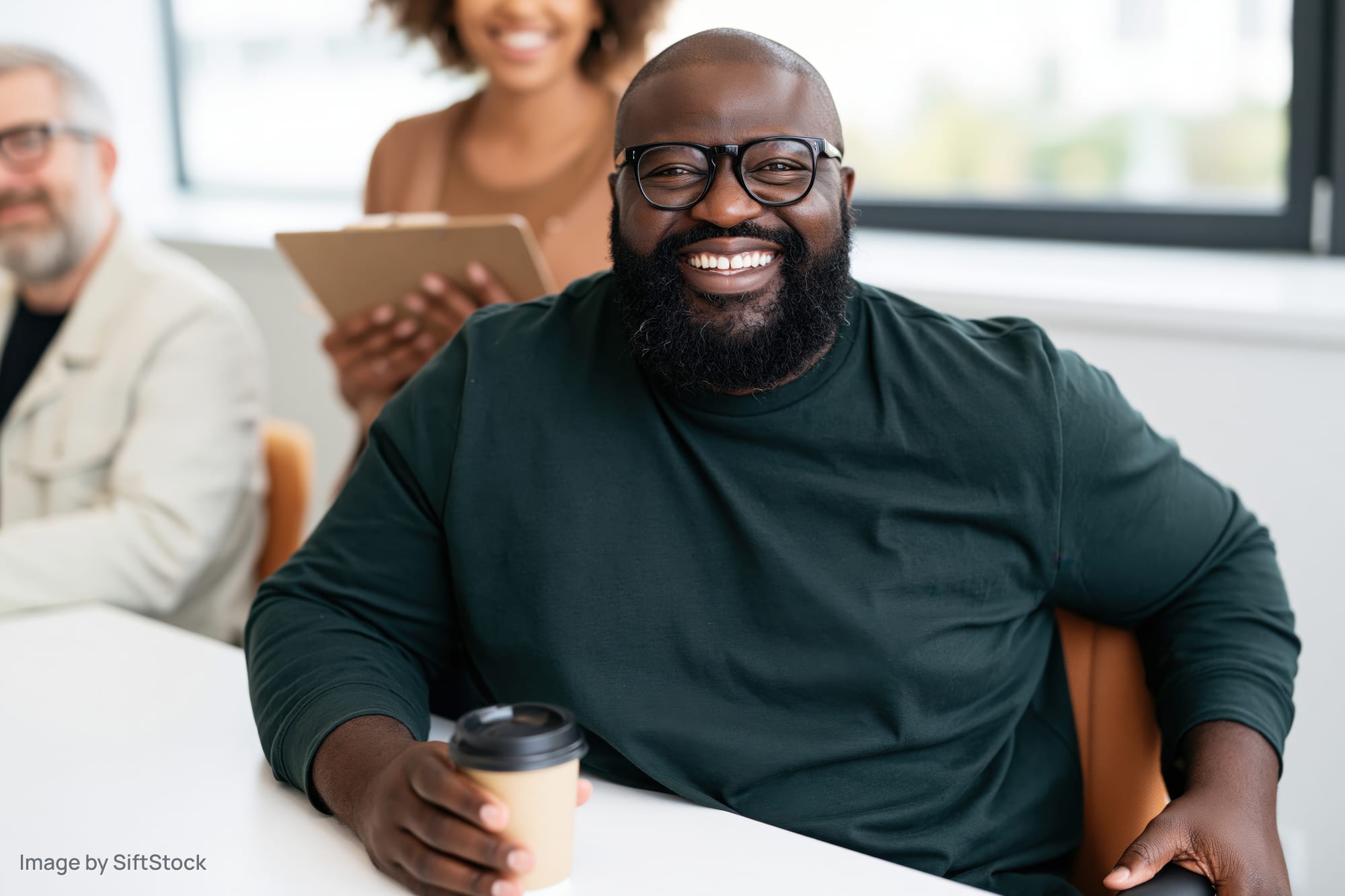 Bearded man with glasses smiling while holding coffee cup at white desk in office meeting with colleagues in background