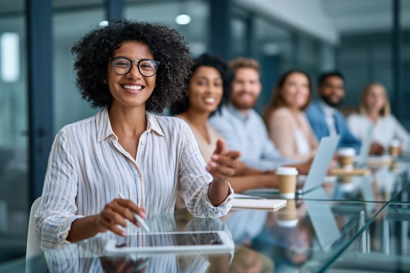 Woman smiling at camera during team meeting in modern office with colleagues at glass desk using tablets and laptops