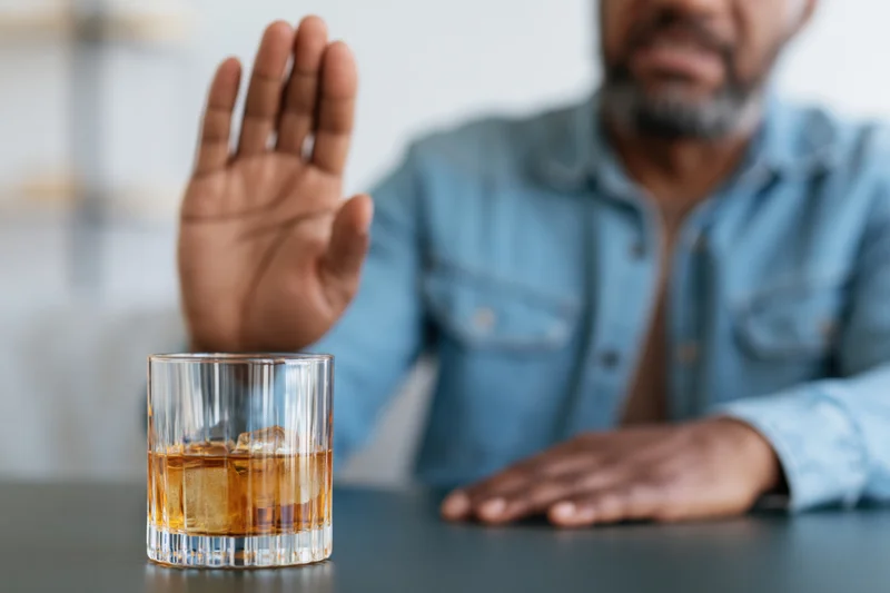 Man raising hand in refusal toward whiskey glass on dark table
