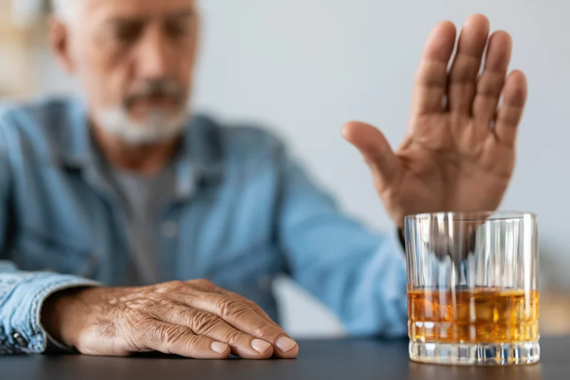 Senior man raising hand in refusal near whiskey glass on dark table