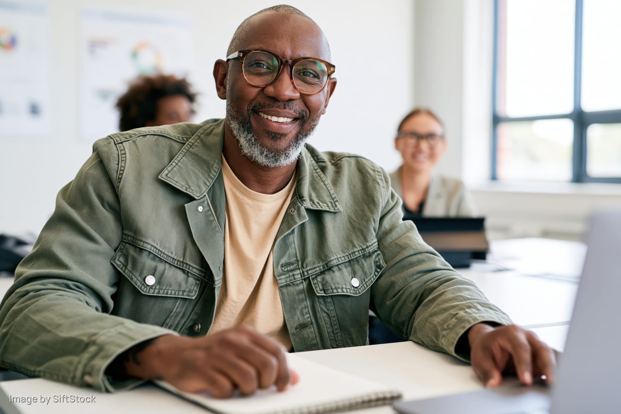 Smiling man with glasses in green denim jacket and beige t-shirt at office desk with laptop and notebook, colleagues in background