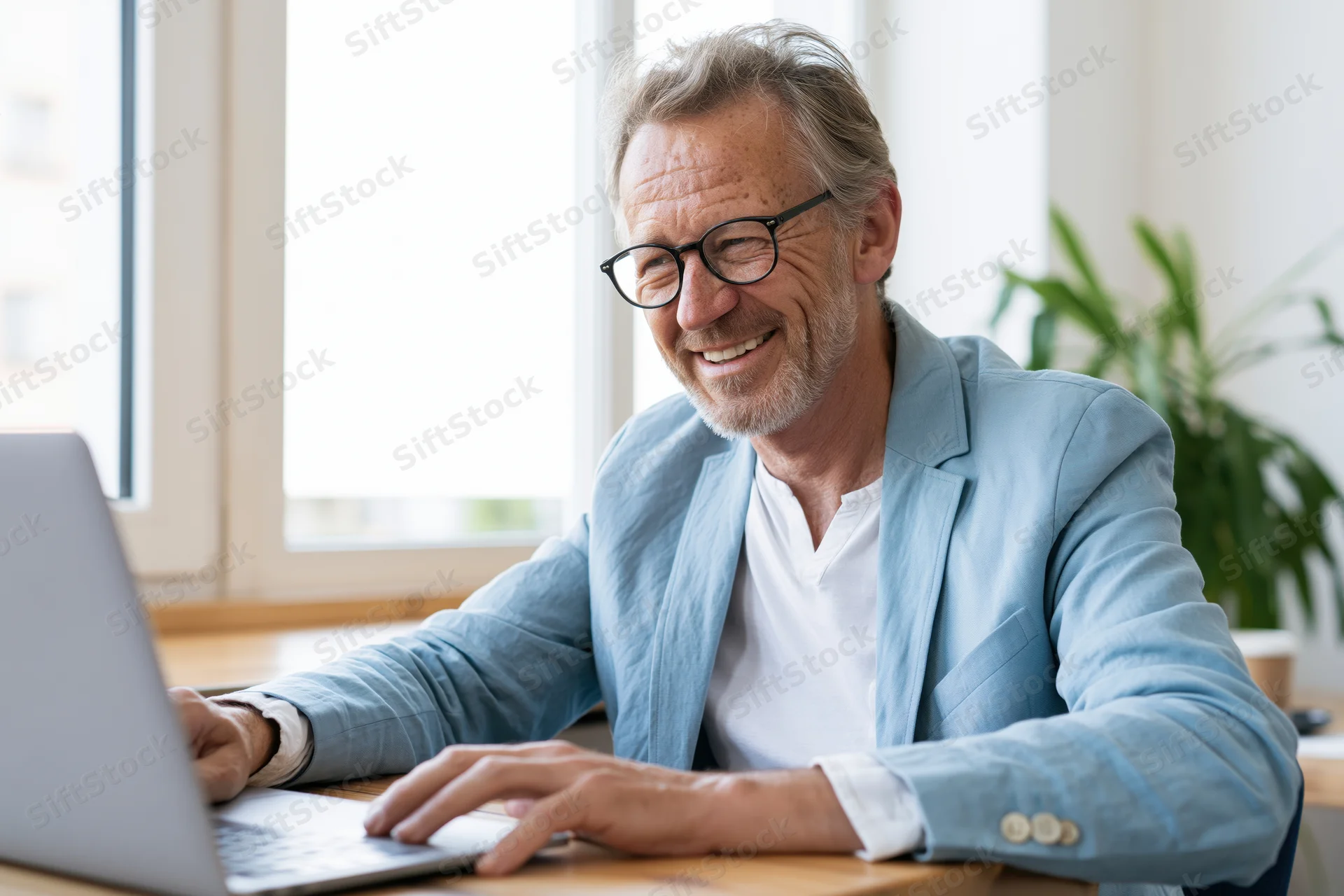 Smiling senior man in glasses working on laptop at desk by window