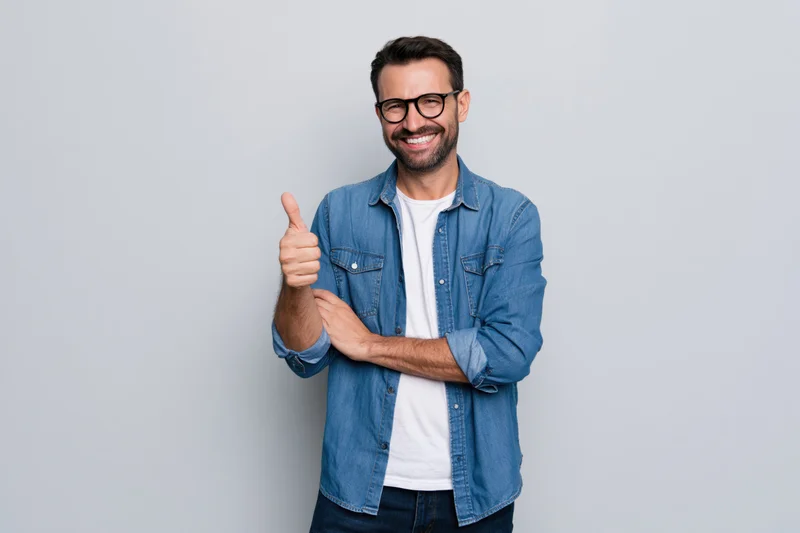 Man smiling and giving thumbs up in denim shirt against light gray background