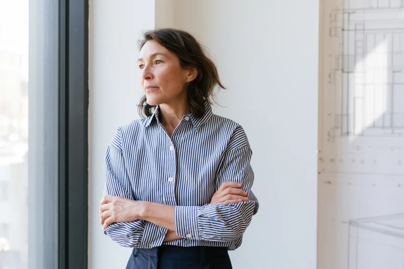 Woman with arms crossed standing by window in office with blueprint on wall