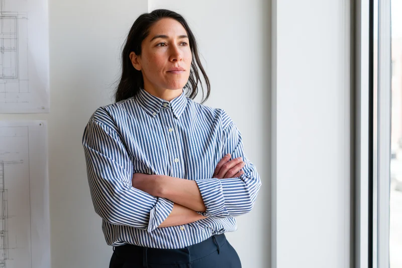 Woman with arms crossed standing by window in office, looking thoughtful near architectural blueprints