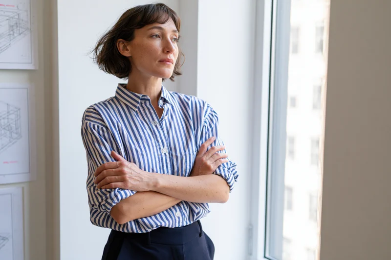 Woman standing by window with arms crossed in office workspace