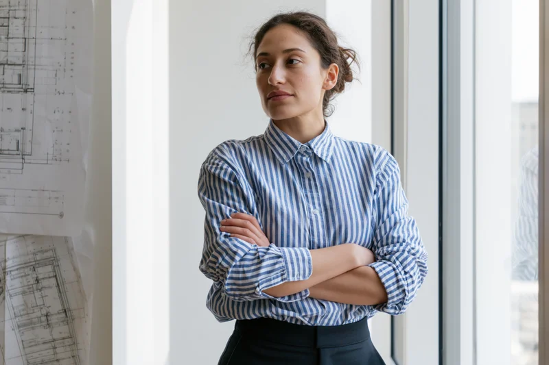 Woman with arms crossed standing by window in office near blueprints