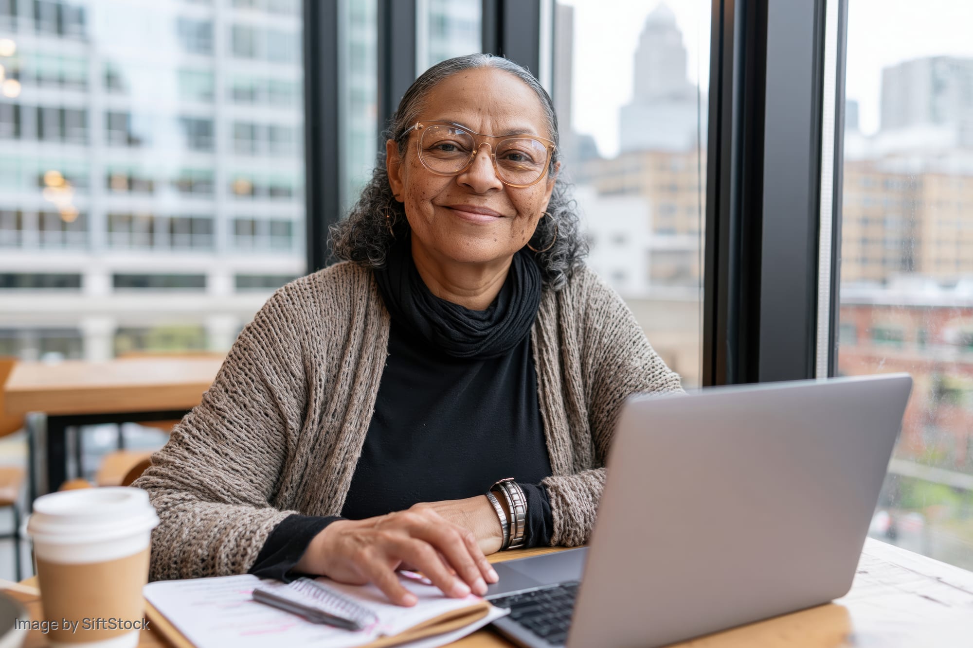 Woman smiling at camera while working on laptop at desk by window in office