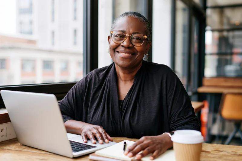 Smiling black senior woman with glasses sitting at wooden desk using laptop and notebook in modern office space near window