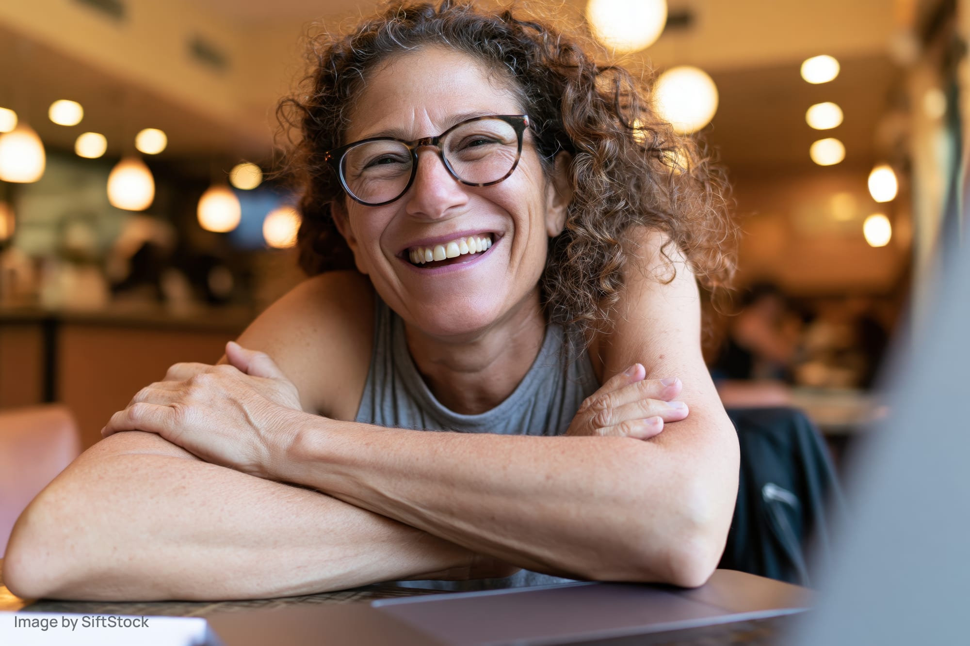 Woman smiling with arms crossed at cafe table near laptop