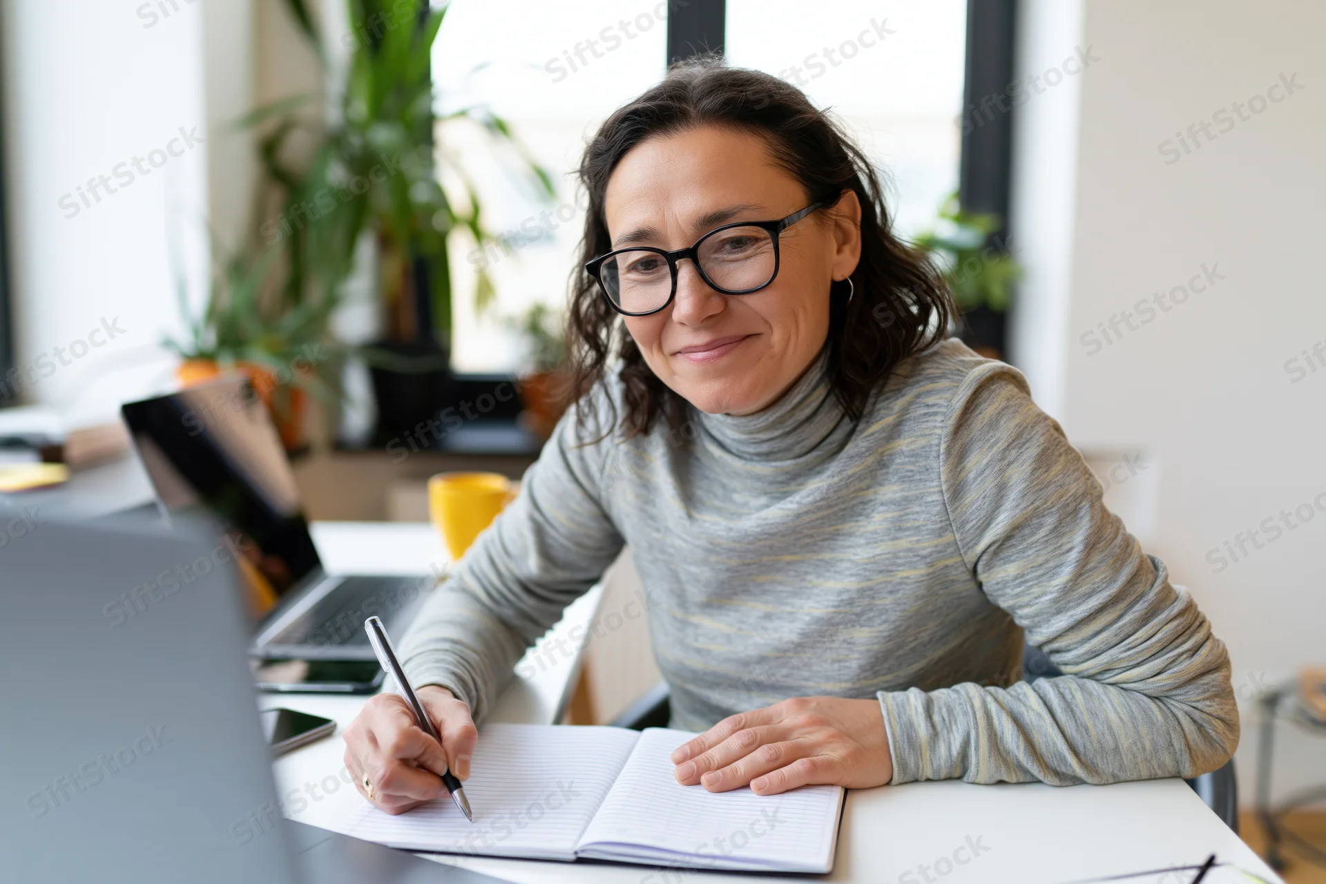 Woman writing in notebook at white desk with laptop and plant in background