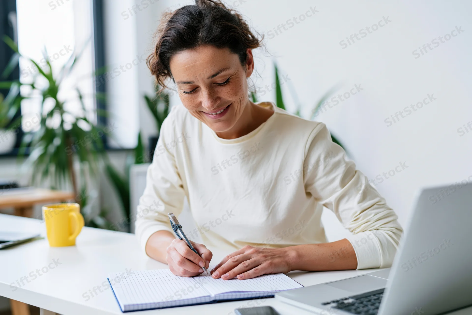 Woman writing in a notebook at a white desk with laptop and mug nearby