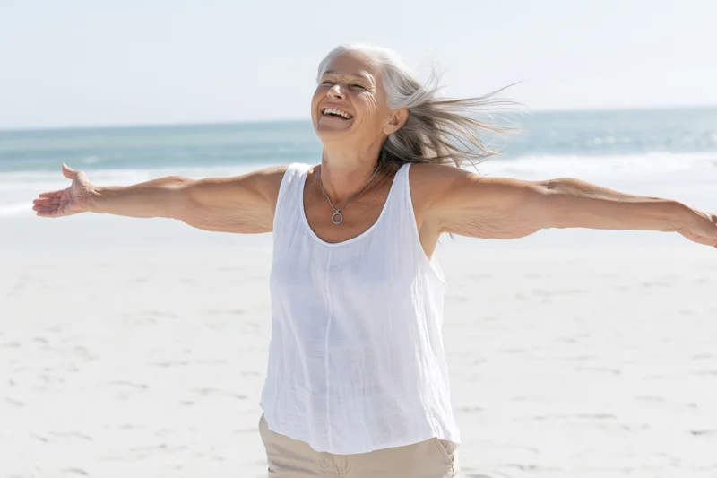 Senior woman laughing with arms outstretched on a sunny beach