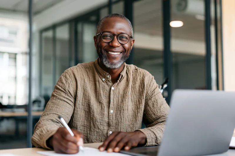Smiling man writing with a pen at a desk in an office with a laptop in front of him