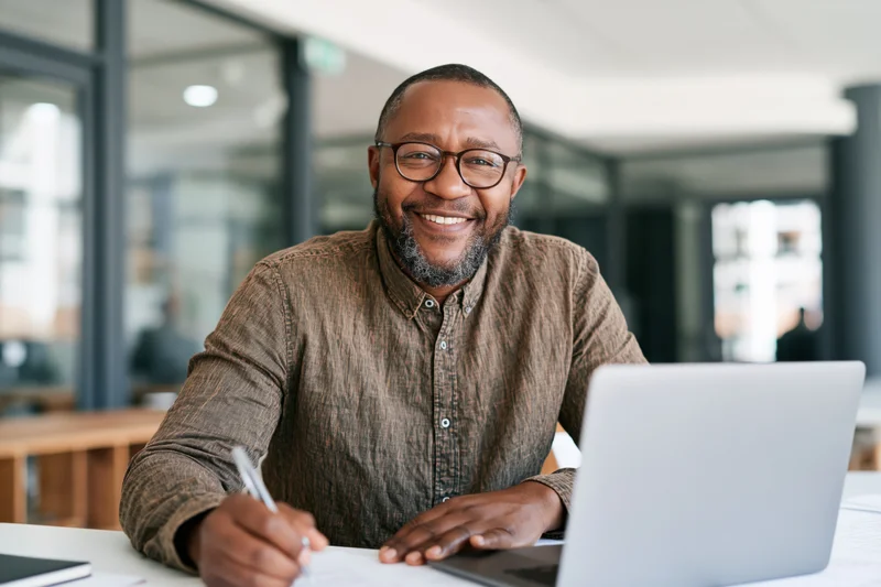 Smiling man writing with pen at desk beside laptop in office
