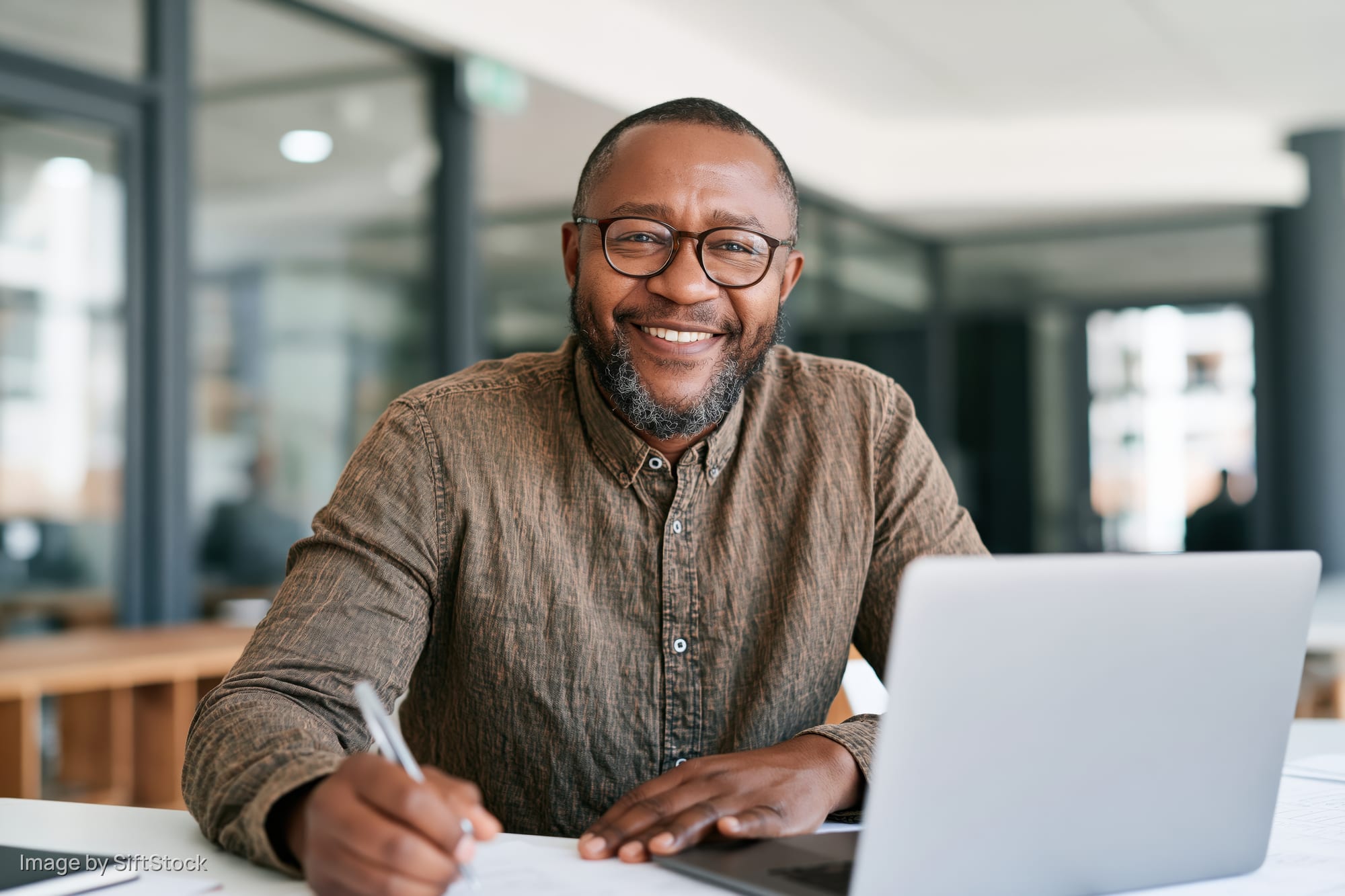 Smiling man writing with pen at desk beside laptop in office