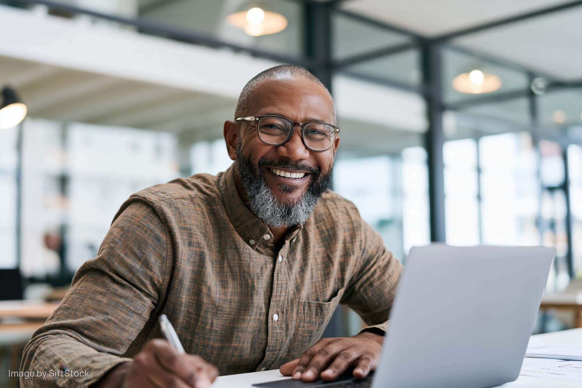 Smiling man writing with pen at desk in modern office with laptop