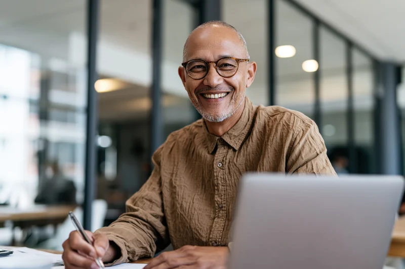 Smiling man writing with pen at desk in modern office with laptop