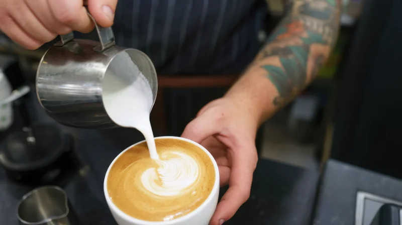 Barista pouring steamed milk into a latte to create foam art in a coffee shop