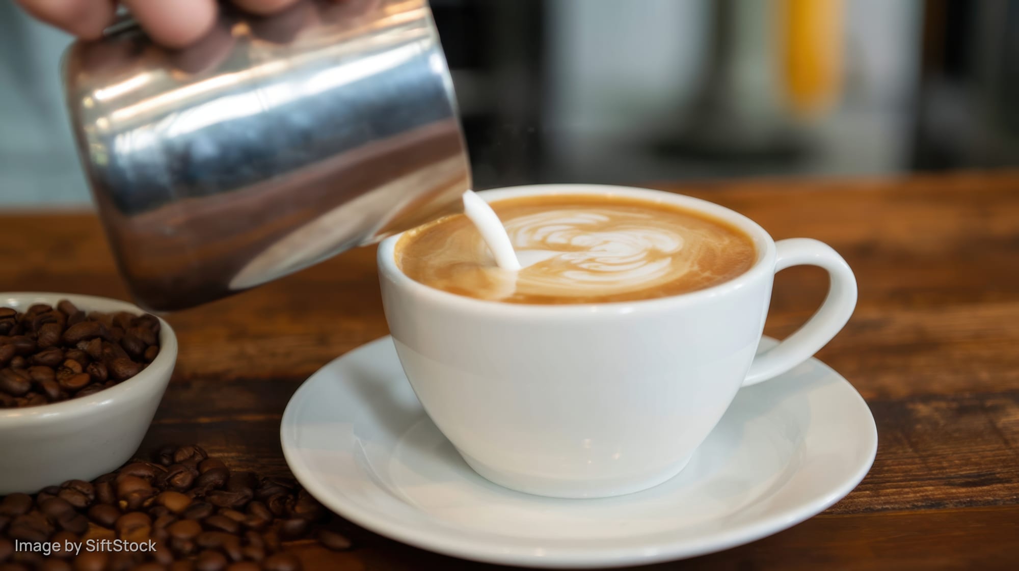 Barista pouring steamed milk into a latte on a wooden table beside coffee beans