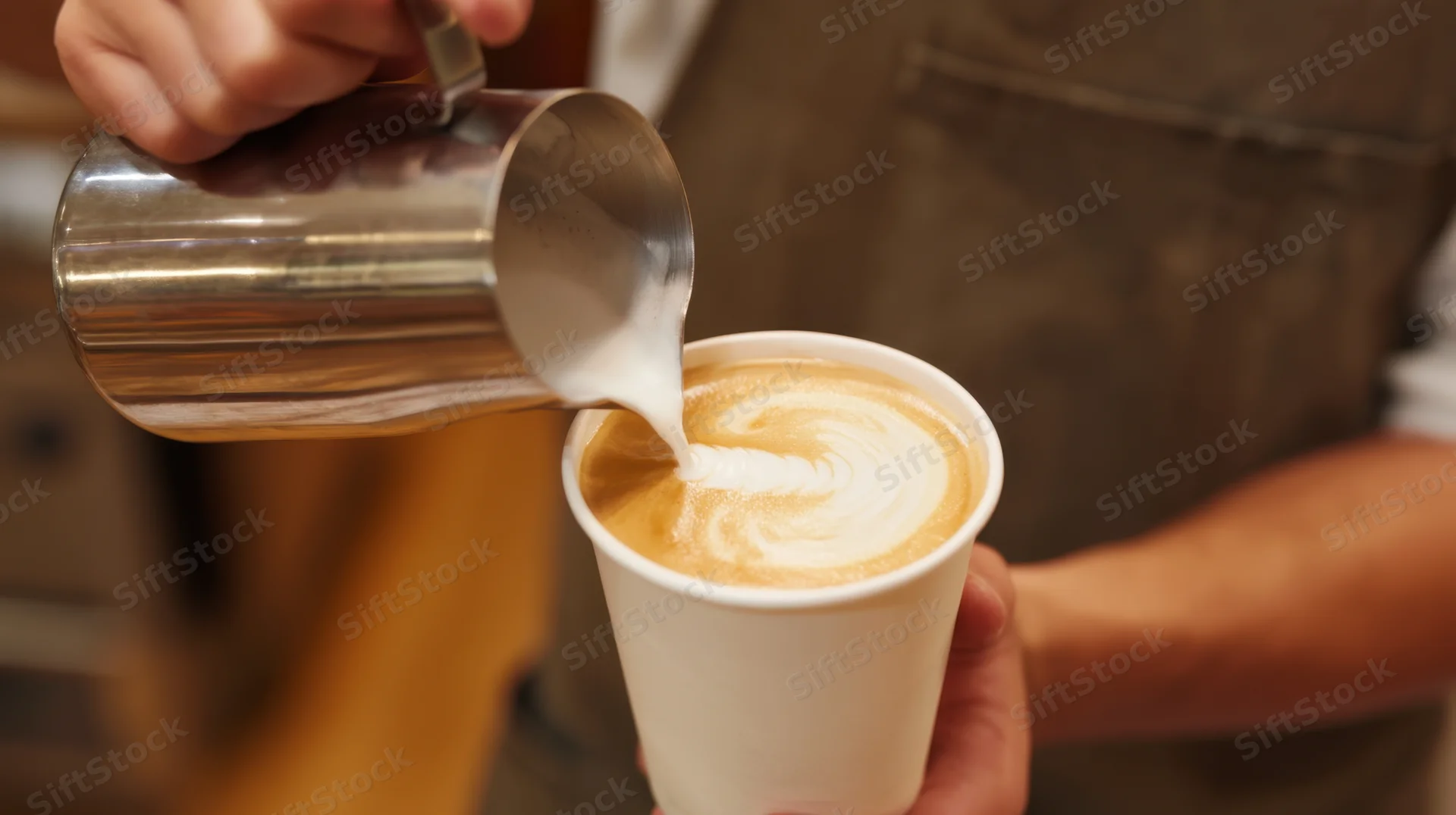 Barista pouring steamed milk into a paper cup of coffee to create latte art in a cafe