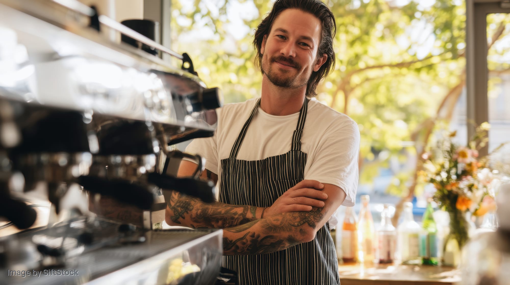Barista with tattoos standing behind coffee machine in cafe with arms crossed and smiling