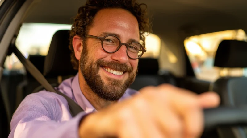 Smiling man with beard and glasses driving a car, hand on steering wheel, wearing purple shirt and seatbelt