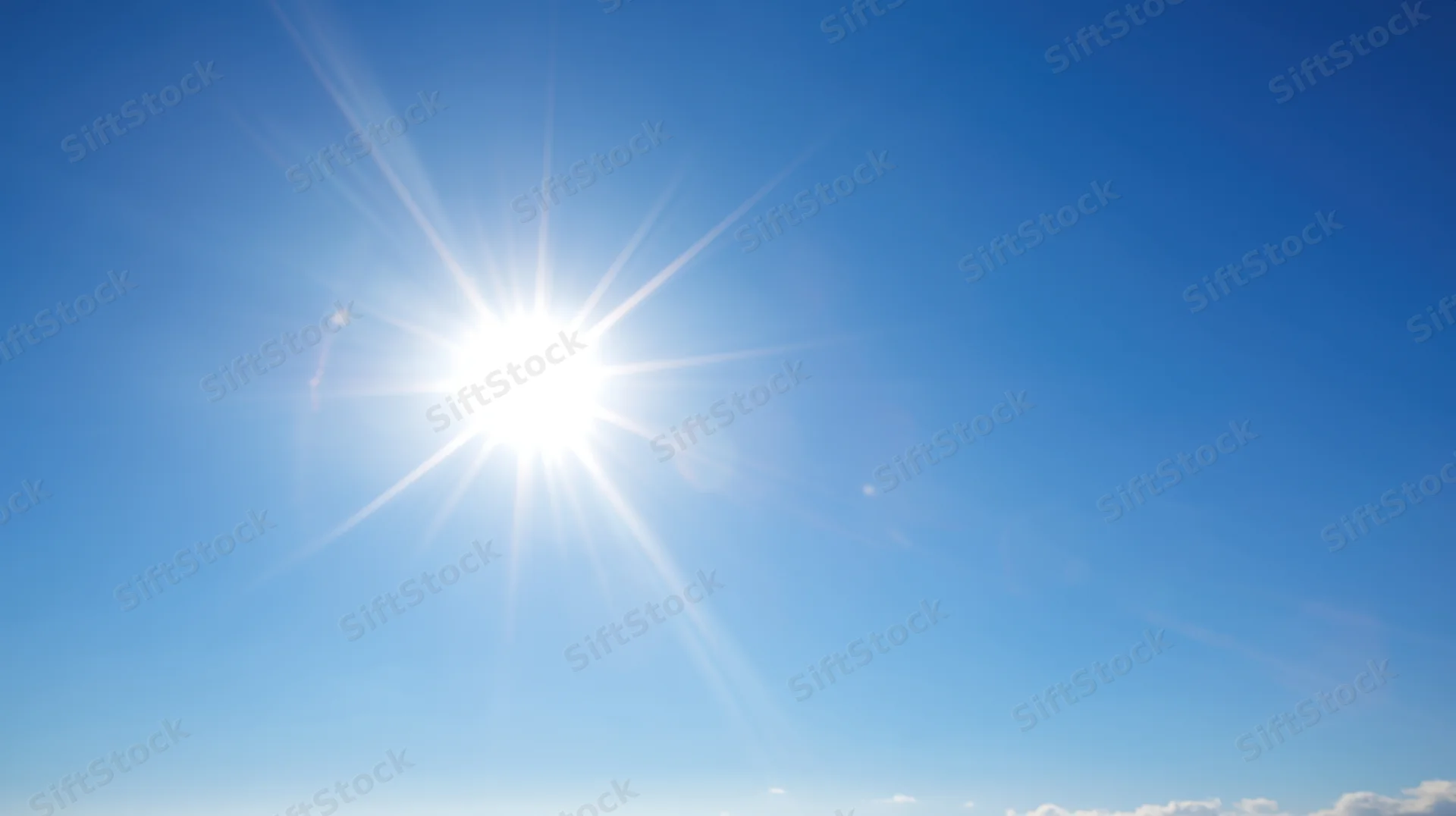 Bright sun shining in clear blue sky with lens flare and scattered clouds at horizon