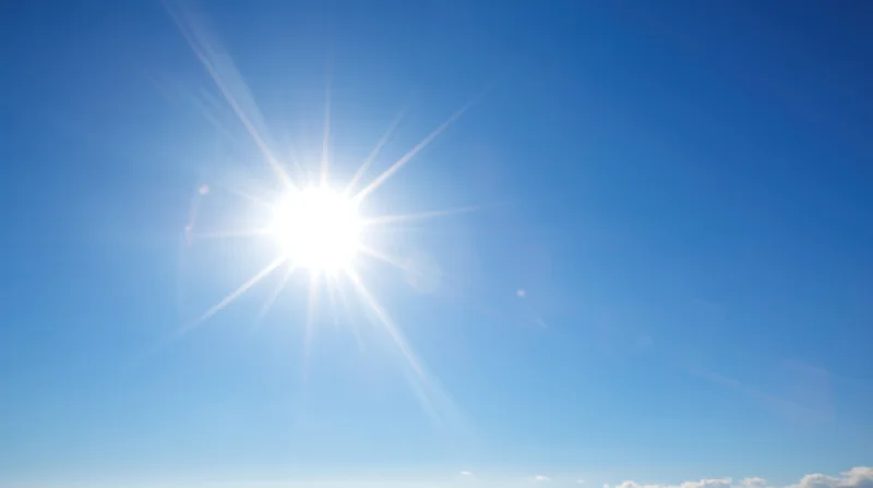 Bright sun shining in clear blue sky with lens flare and scattered clouds at horizon