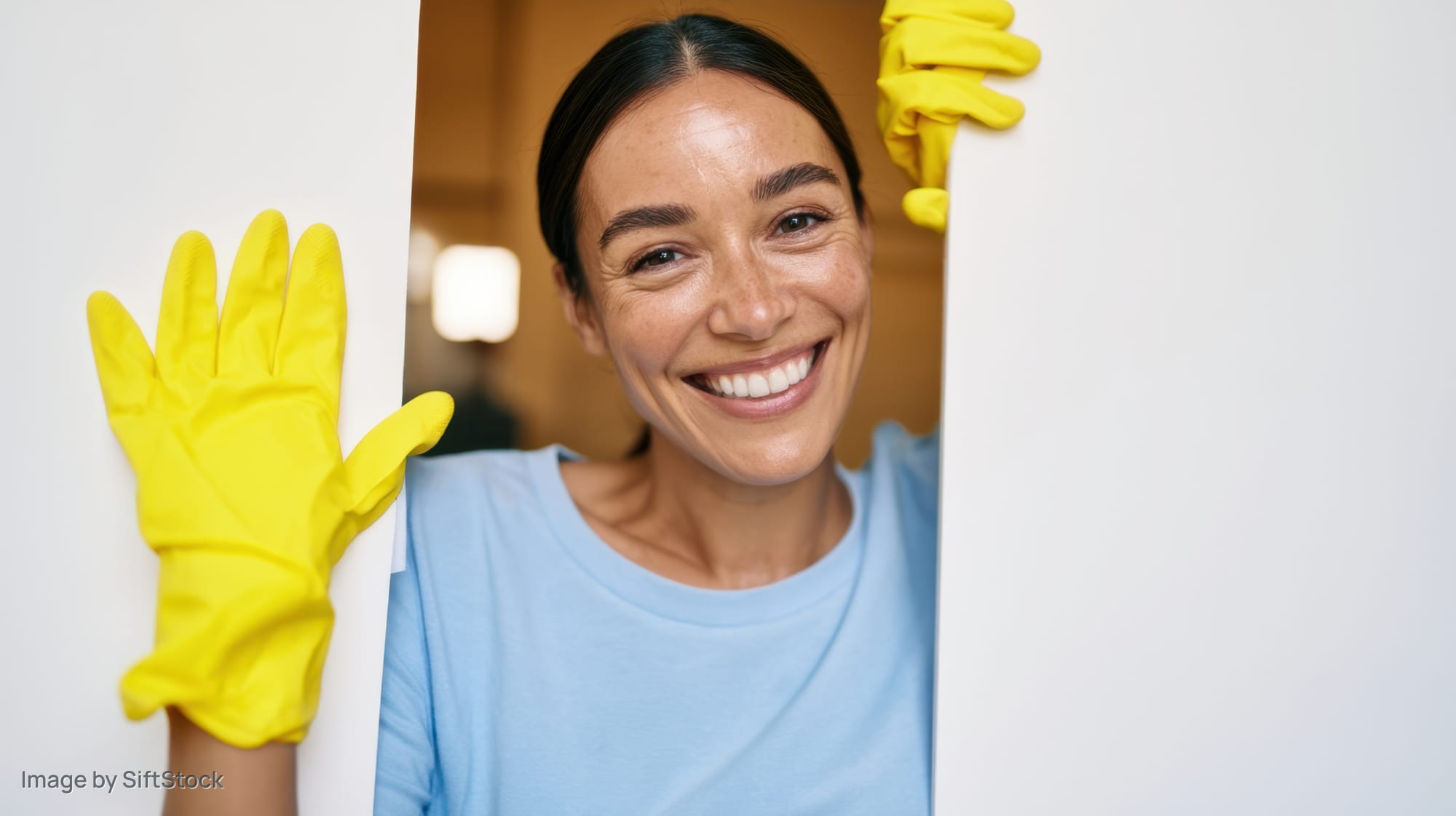 Woman smiling while peeking from behind a white panel wearing yellow cleaning gloves