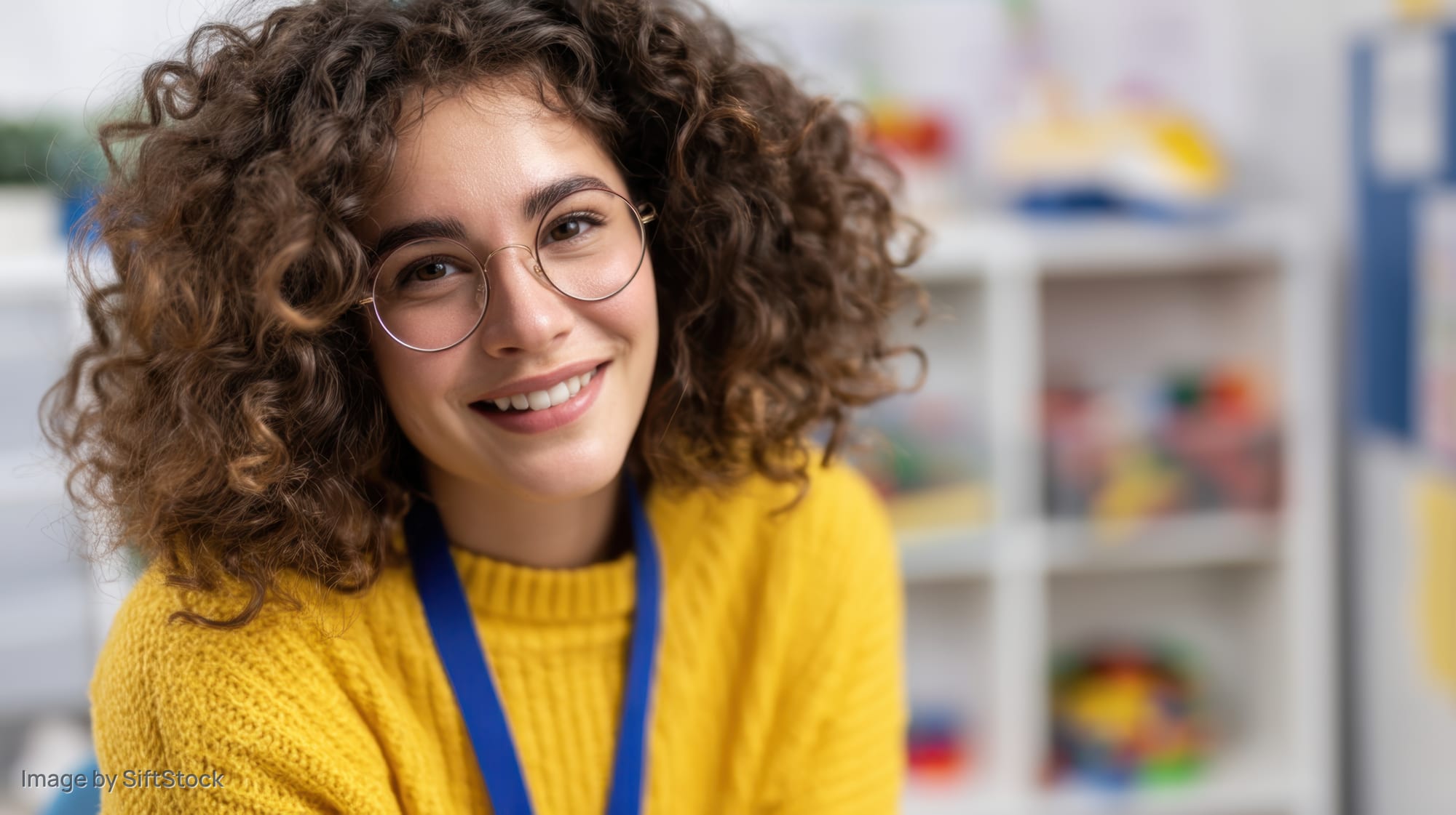Smiling woman with curly hair and glasses wearing yellow sweater and blue lanyard in classroom setting