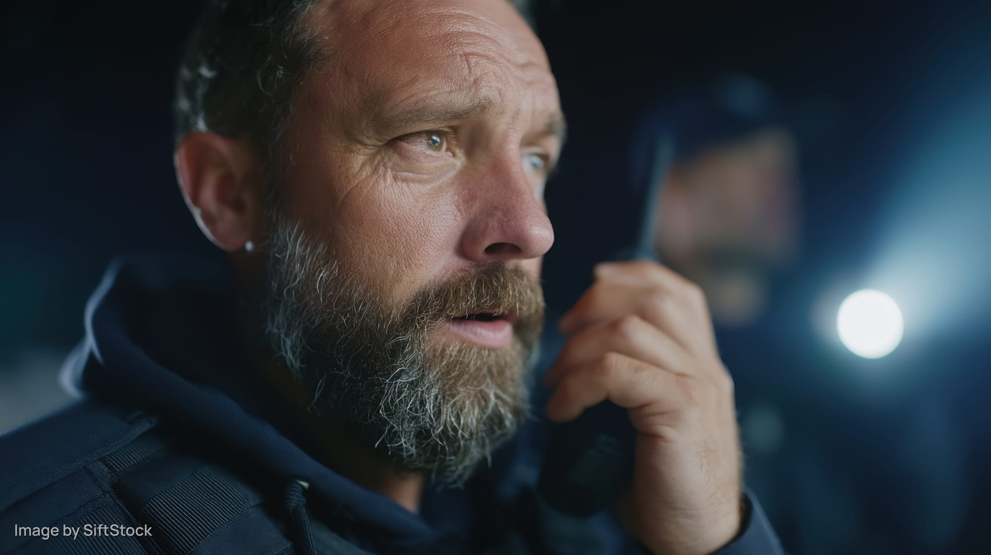 Bearded man in uniform holding radio at night with flashlight in background