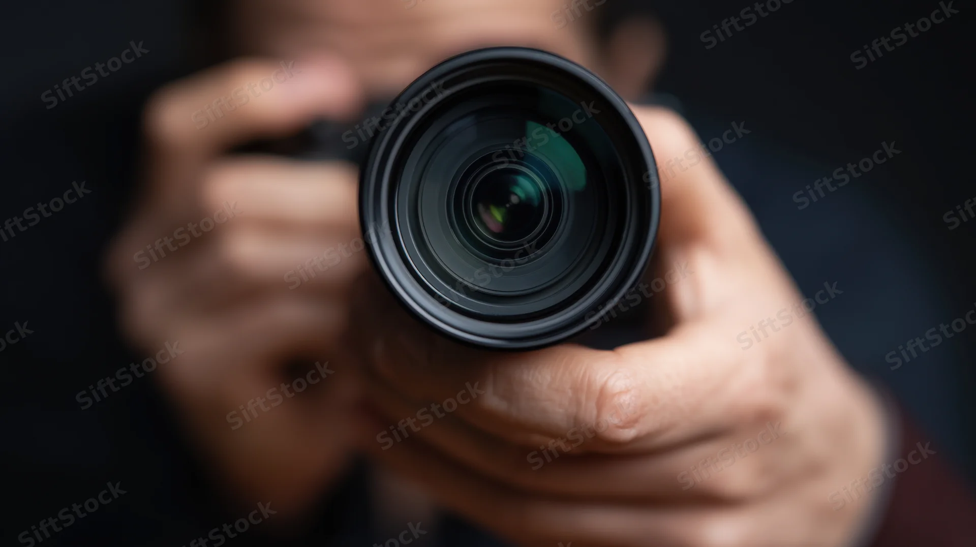 Close-up of a camera lens held by hands in focus against a dark blurred background