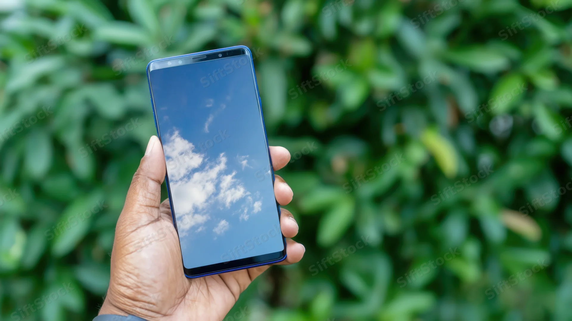 Hand holding smartphone with sky and clouds reflected on screen outdoors near green foliage