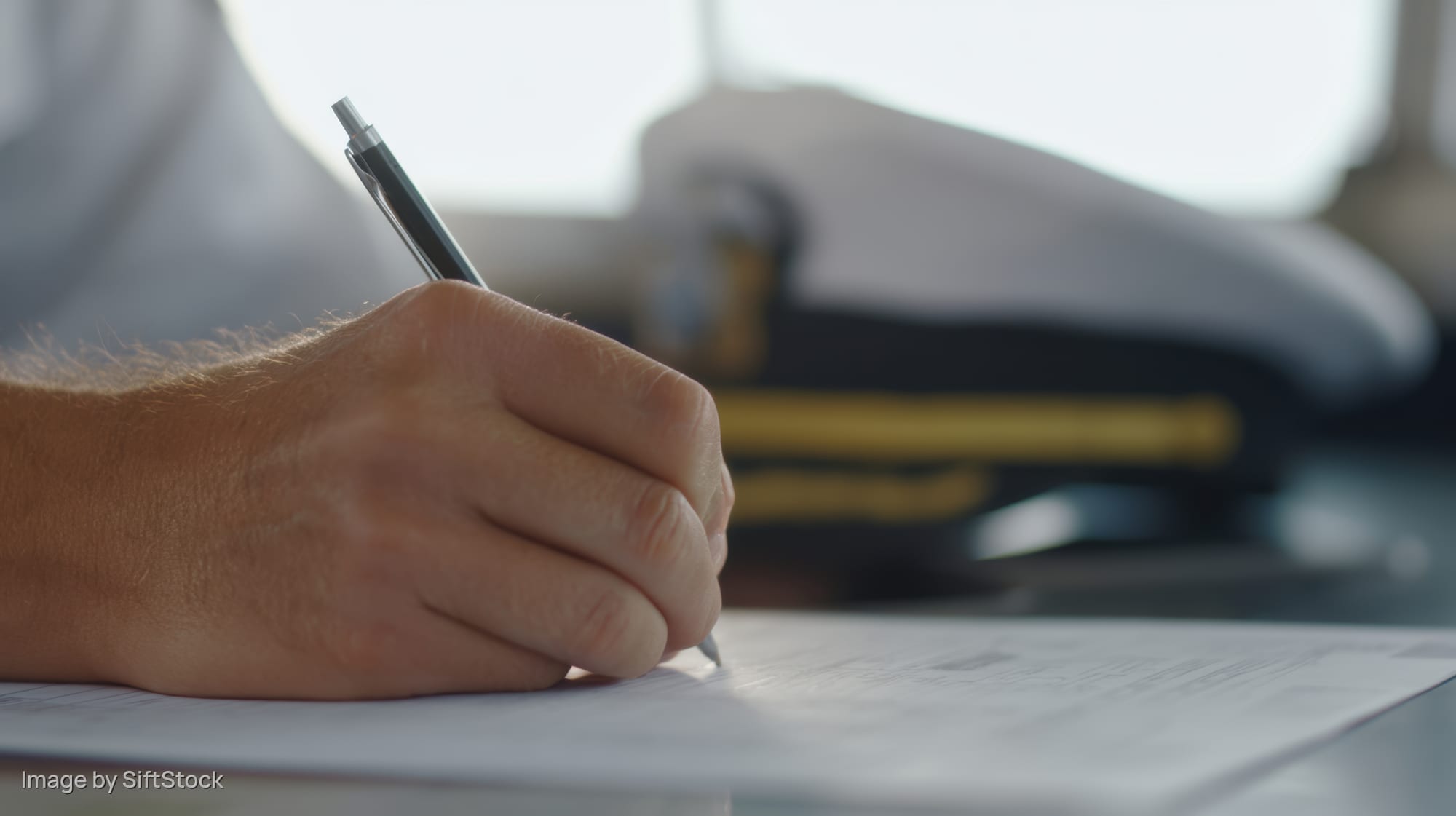 Hand writing with pen on document at desk with pilot cap in background