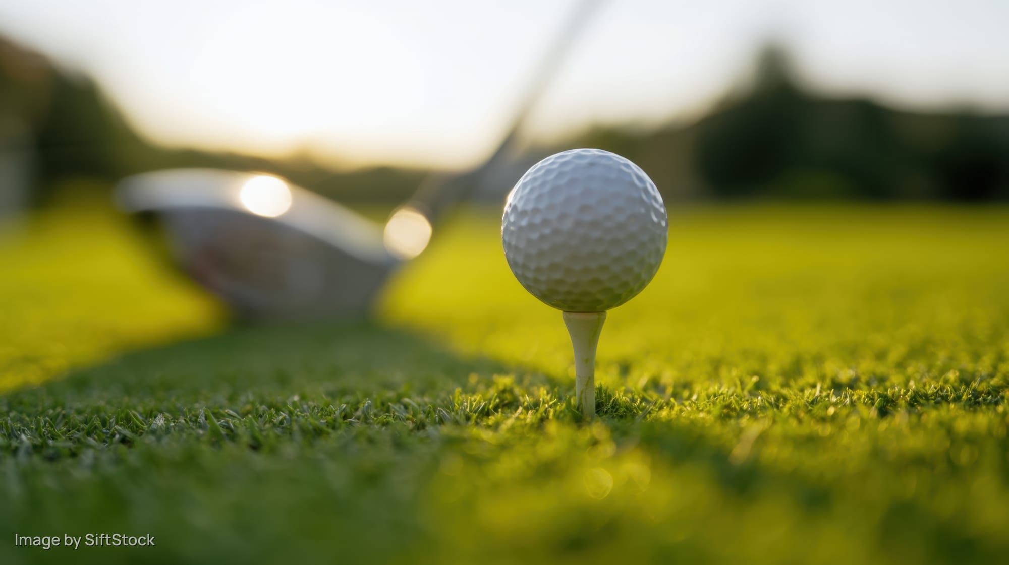 Golfball on tee in grass with golf club in background on course