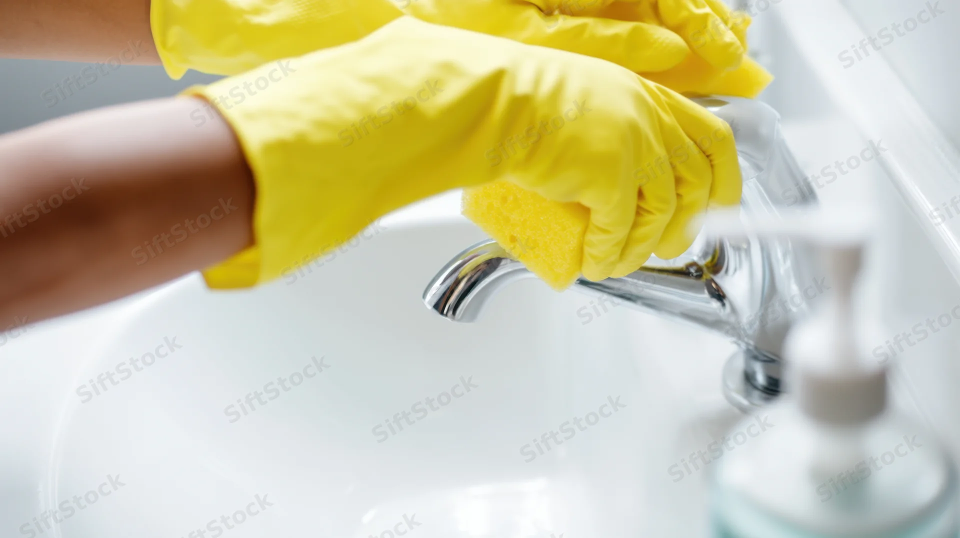 Hands in yellow gloves scrubbing a chrome faucet over a white sink