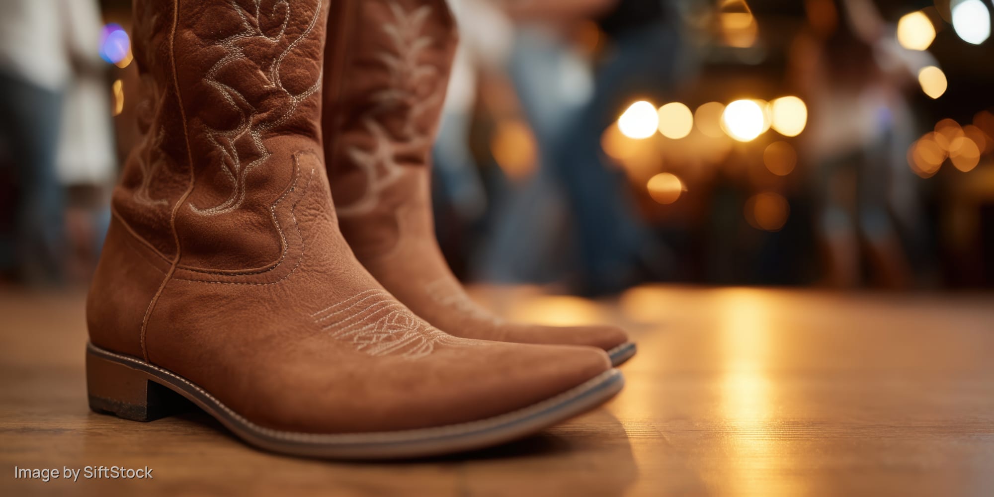 Brown cowboy boots on wooden floor at a lively indoor party with warm bokeh lights in background