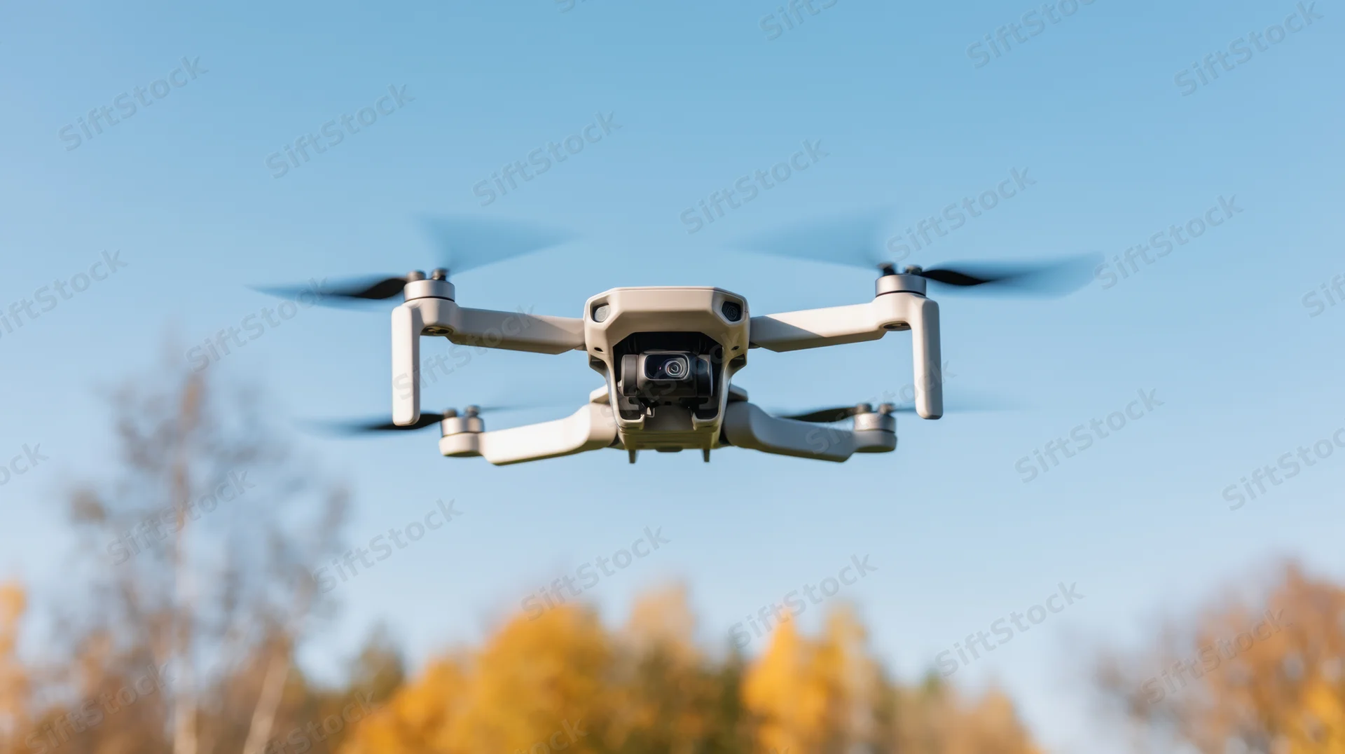 Compact quadcopter drone flying with camera against blue sky above autumn trees