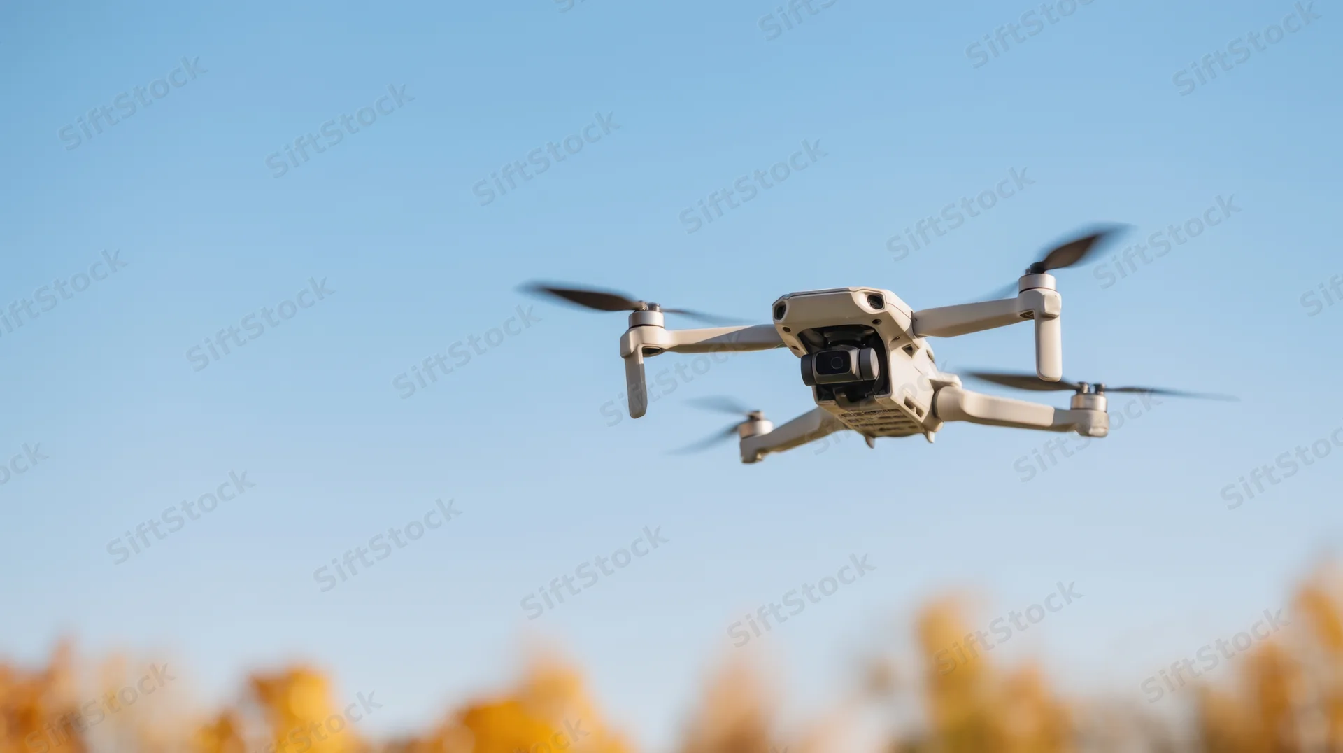 Compact quadcopter drone flying in clear blue sky above autumn foliage