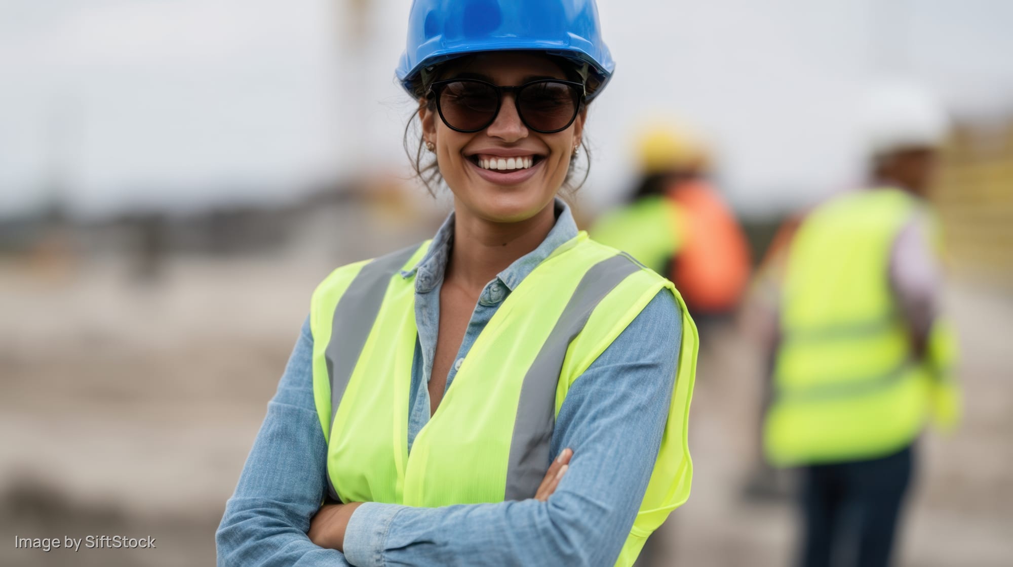 Woman in blue hard hat and yellow safety vest smiling with arms crossed on construction site