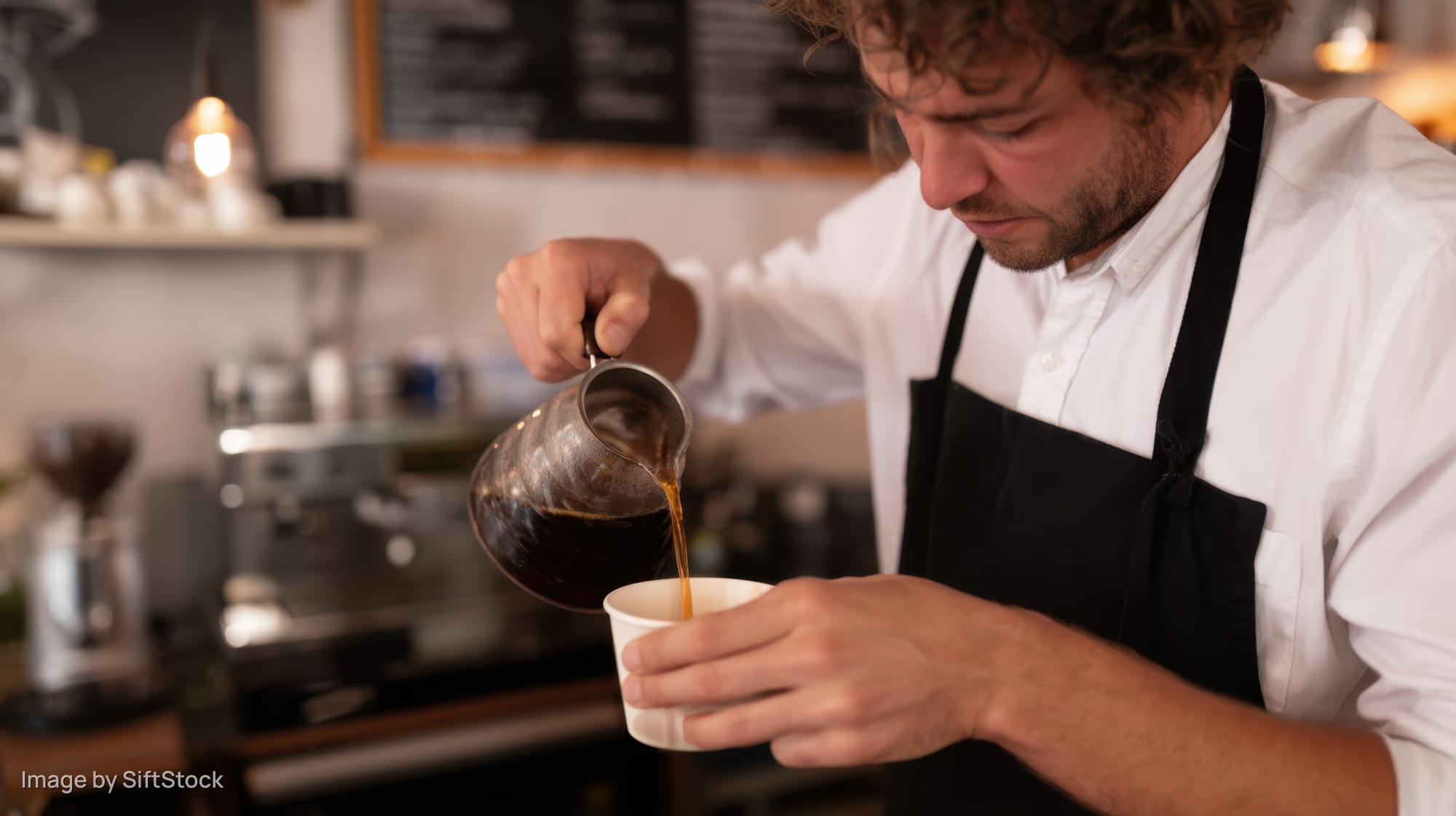 Man in white shirt and black apron pouring coffee from metal pitcher into white cup in cafe