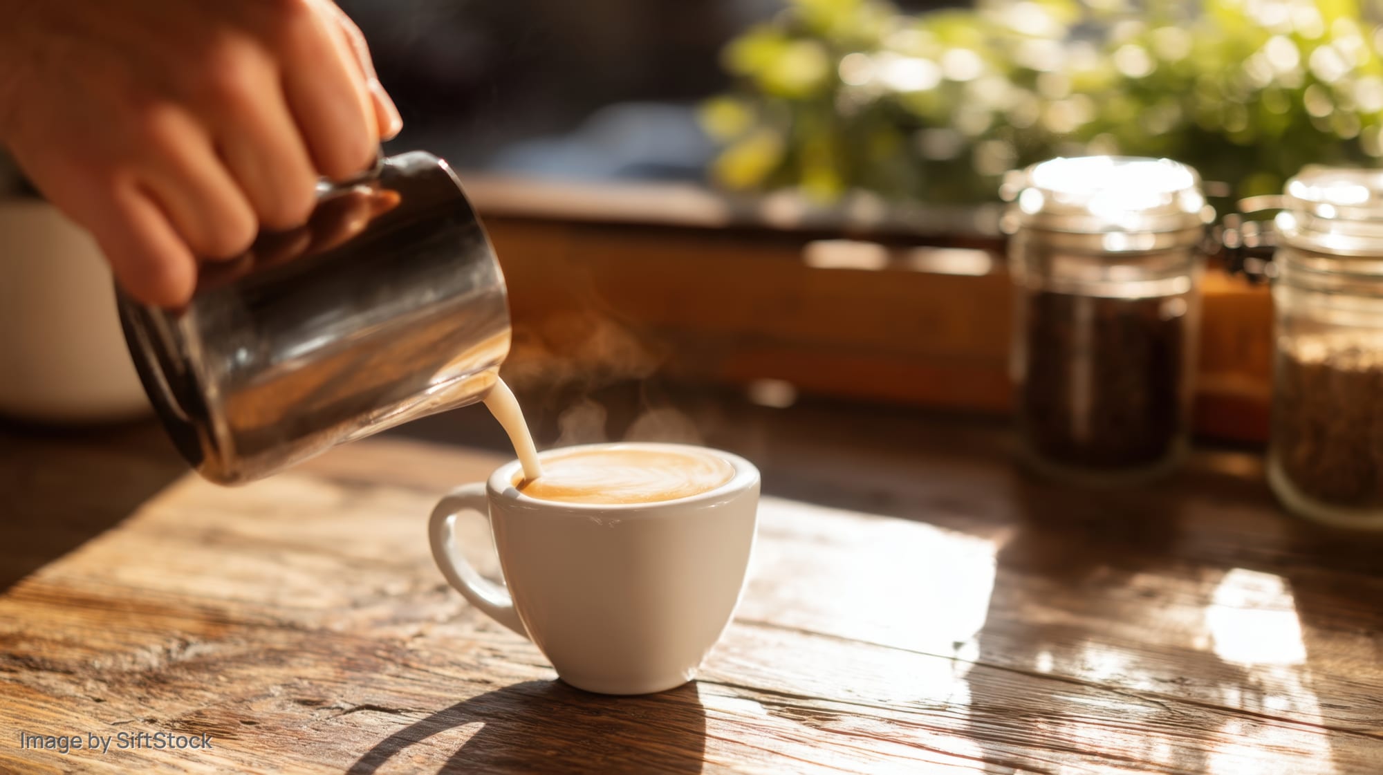 Hand pouring steamed milk into a latte cup on a wooden table in a café