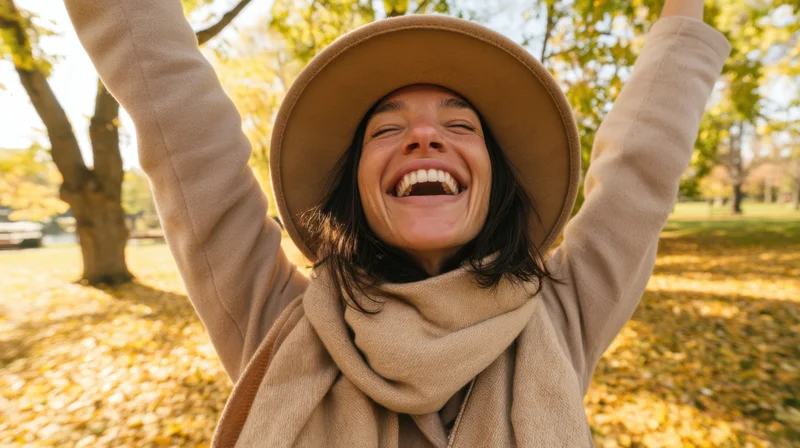 Woman laughing with arms raised in autumn park surrounded by golden leaves